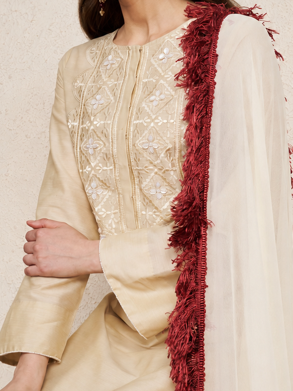 A woman models a traditional Indian outfit featuring a beige kurta with intricate white embroidery on the chest, paired with a cream dupatta draped over her shoulder and a deep red fringed accent, set against a textured beige wall.