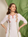 A woman modeling a white cotton kurta with pink floral embroidery and green zigzag border details, posed against a split background of textured beige wall and green fabric.