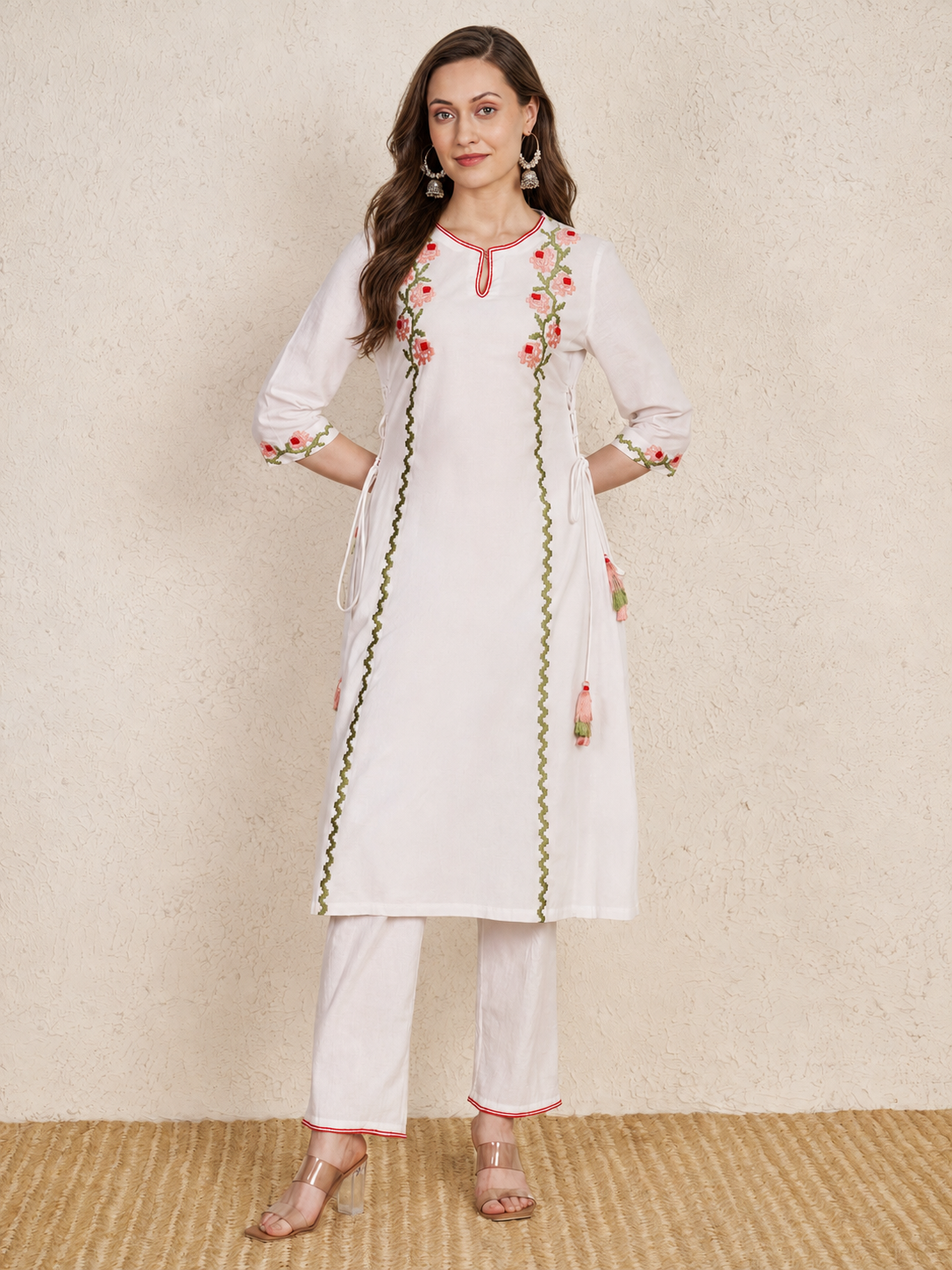 A woman models a white kurta set with pink and green floral embroidery and border details, paired with matching white trousers, against a textured beige wall and woven floor.
