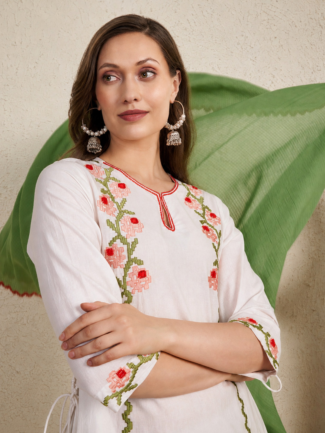 A woman wearing a white kurta with pink and green floral embroidery, paired with a green dupatta, set against a textured beige wall.